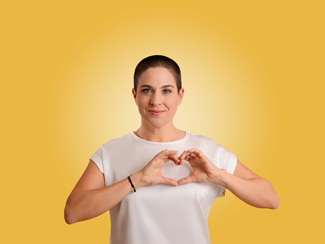 Rebekka in weissem Shirt formt ein Herz mit ihren Händen vor einem gelben Hintergrund – Symbol für Selbstliebe, Achtsamkeit und persönliche Entwicklung im Retreat.